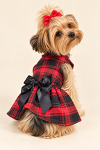 A small Yorkshire Terrier dog wearing a red and black plaid dress with a large black bow, sitting against a plain beige studio background.